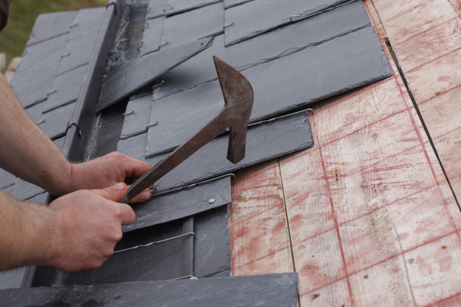 A roofer uses a traditional slate hammer to install dark gray slate tiles on a roof over a wooden sub-roof.