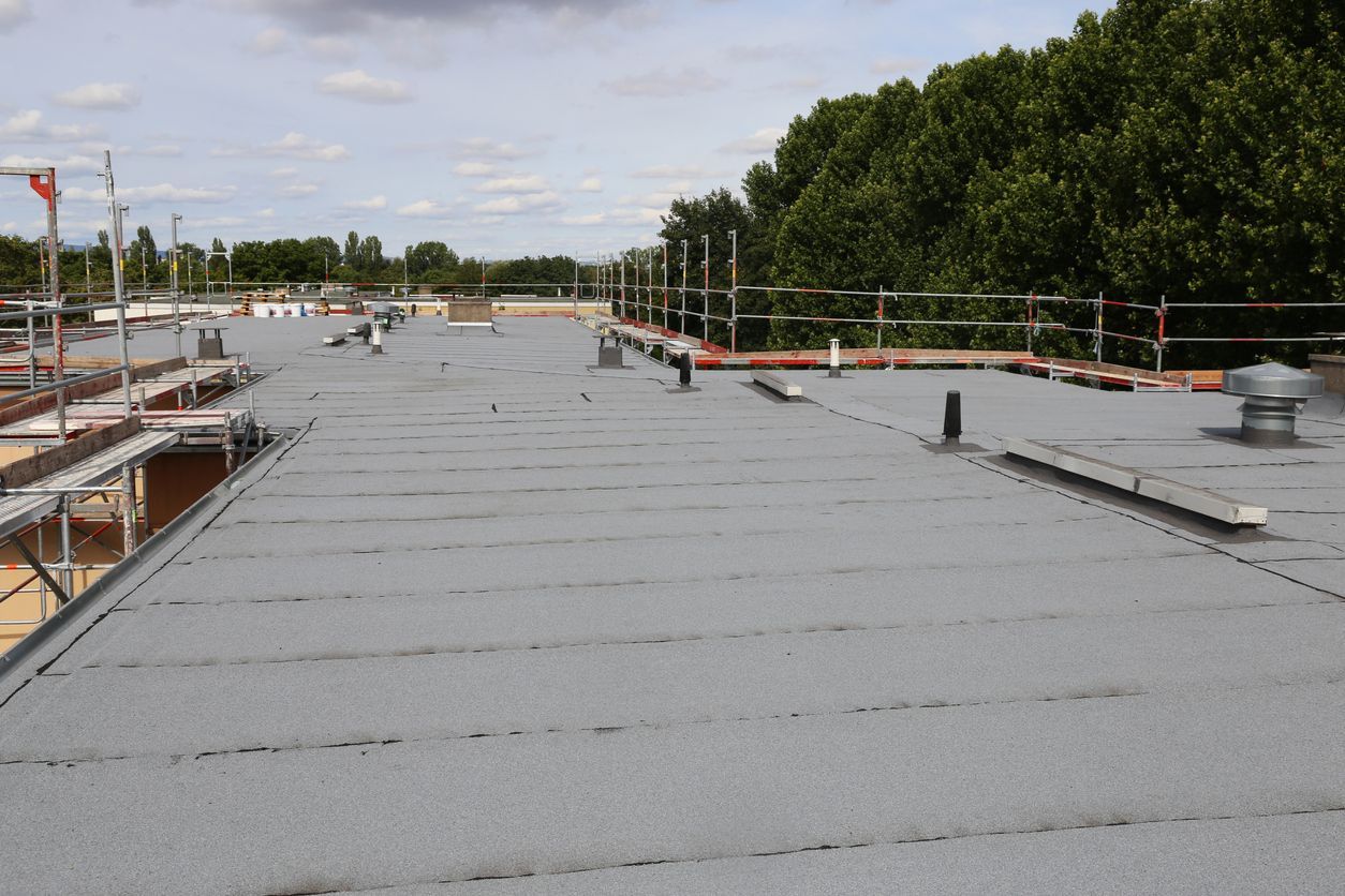 A wide view of a flat roof under construction, featuring gray roofing material, scaffolding, and vent pipes.