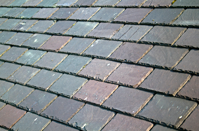 A close-up of a weathered, dark grey slate tile roof with overlapping rows of textured square tiles.