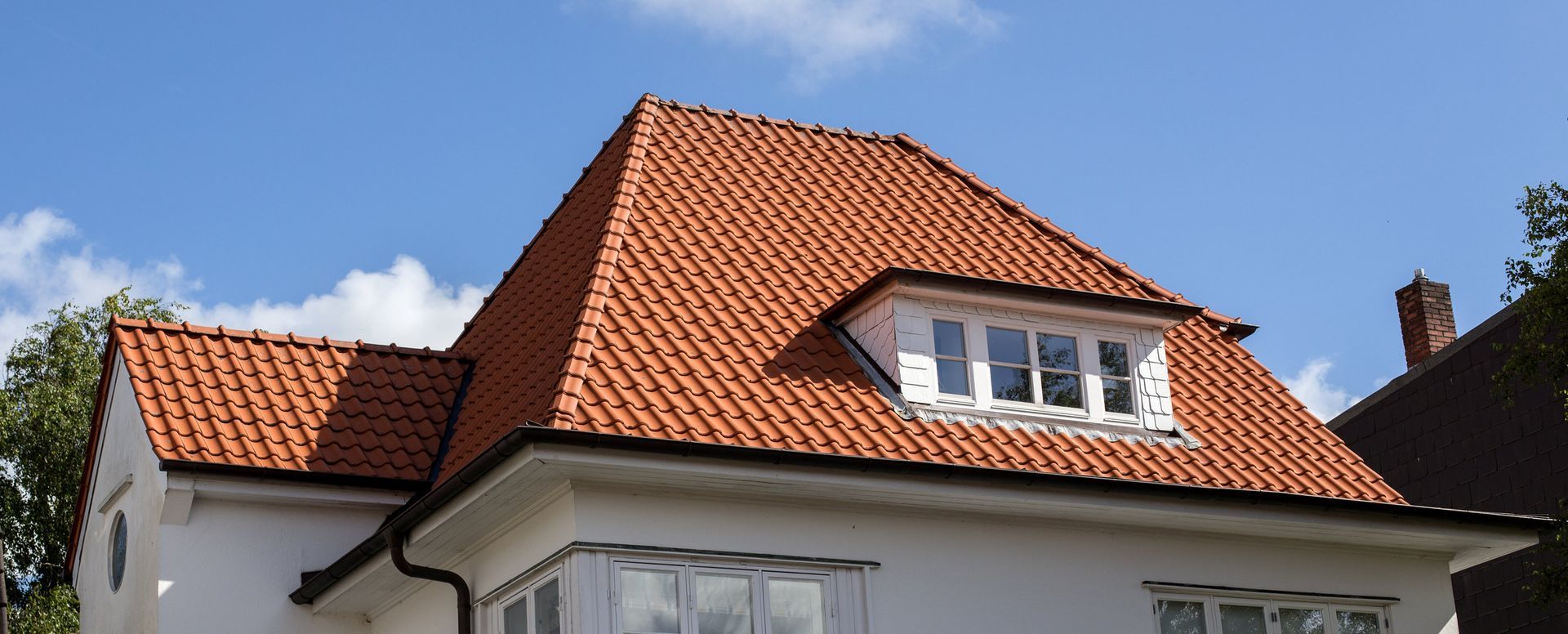 A house roof with red clay tiles, a dormer window, and a white exterior wall against a blue sky with clouds.
