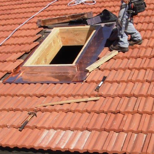 A worker installs a skylight with copper flashing on a roof covered in red clay tiles.