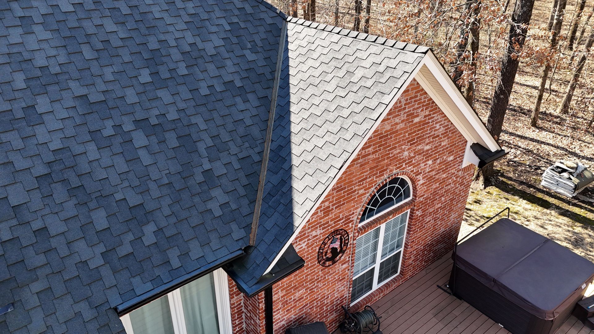 An aerial view of a brick house with a partially shingled roof, featuring a dark blue section next to a grey section.