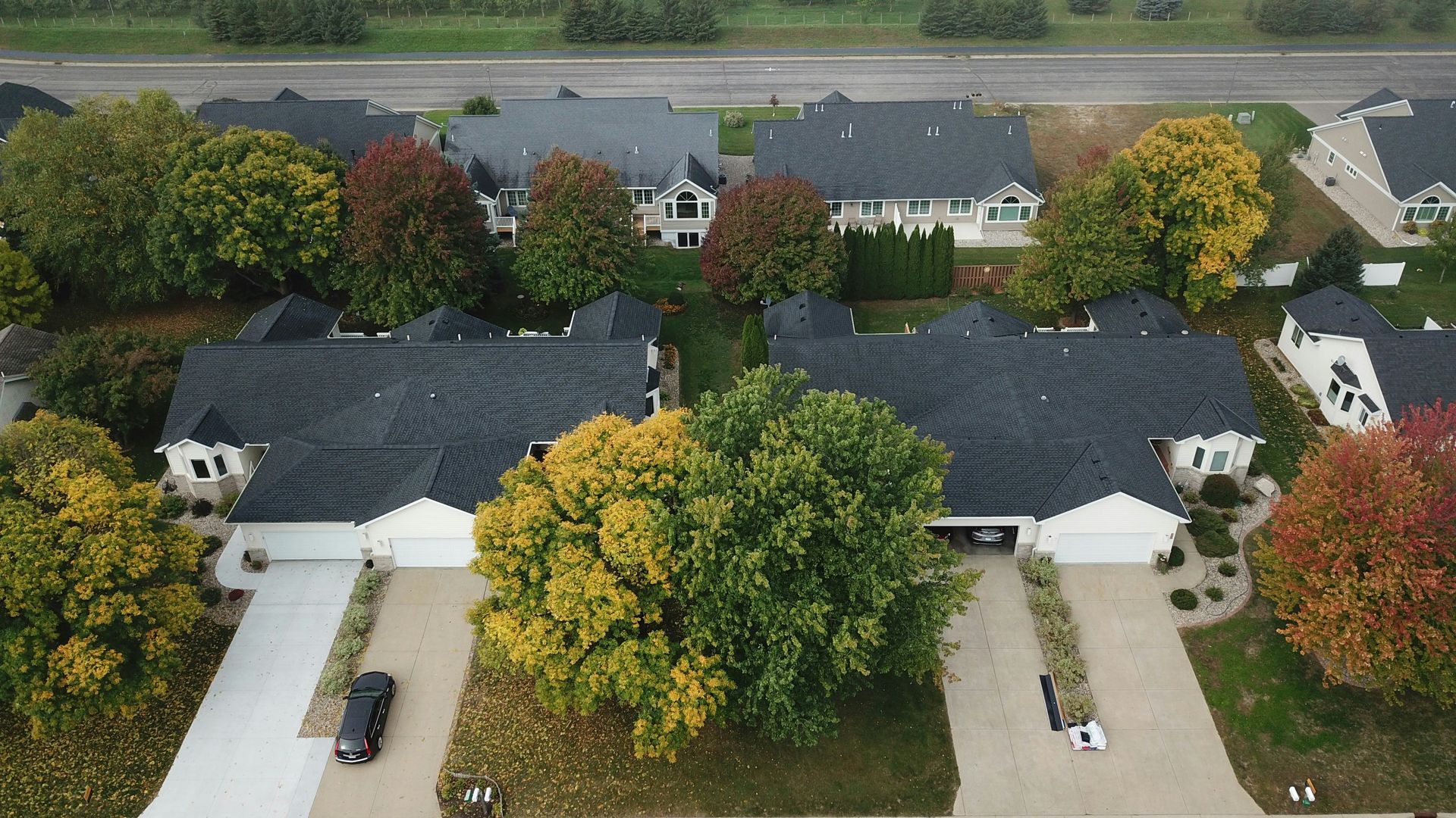 An aerial view of suburban duplexes with dark roofs, concrete driveways, and surrounding trees showing autumn colors.