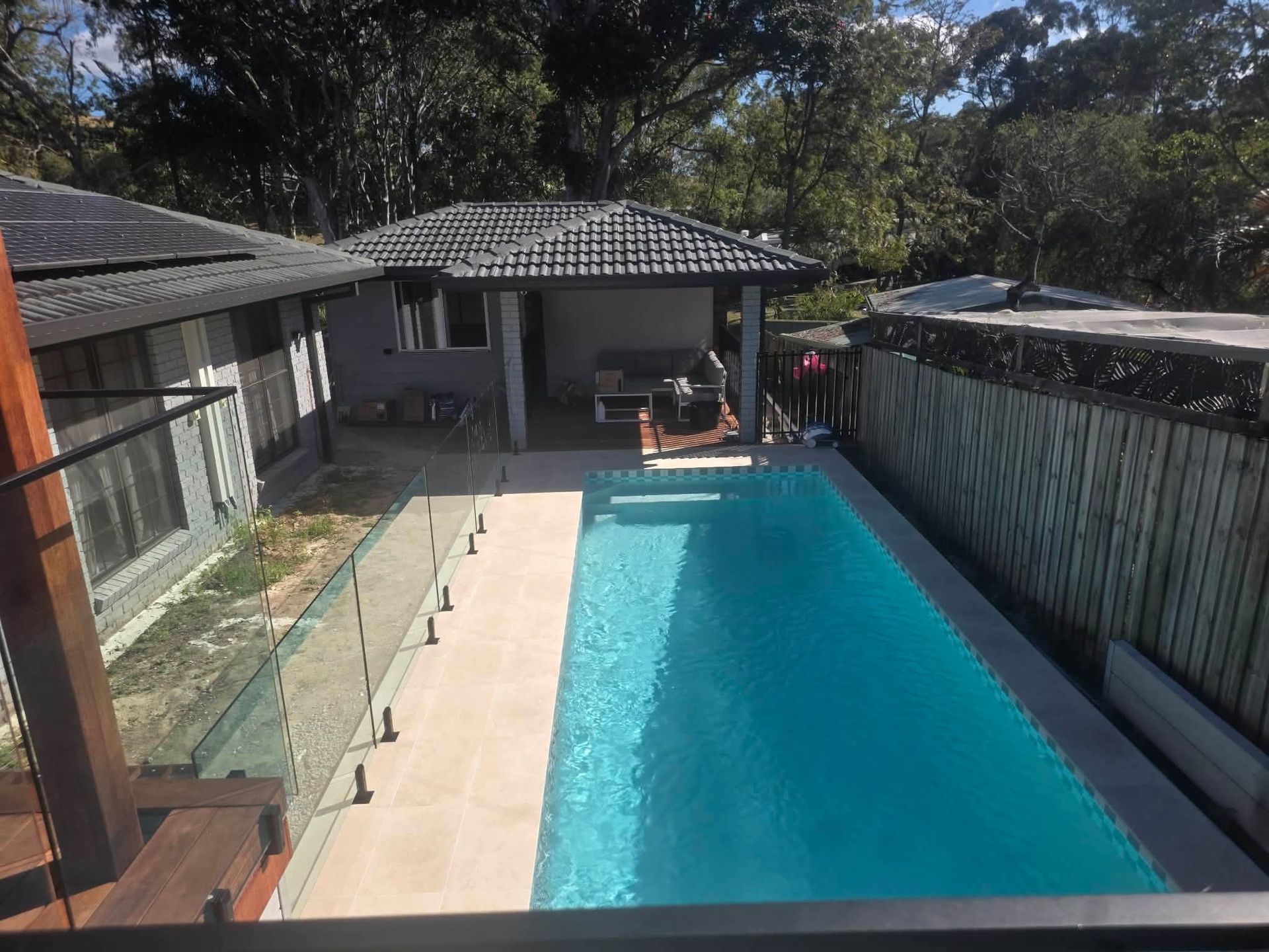 Backyard swimming pool beside a patio and house, with glass fence and tiled roof.