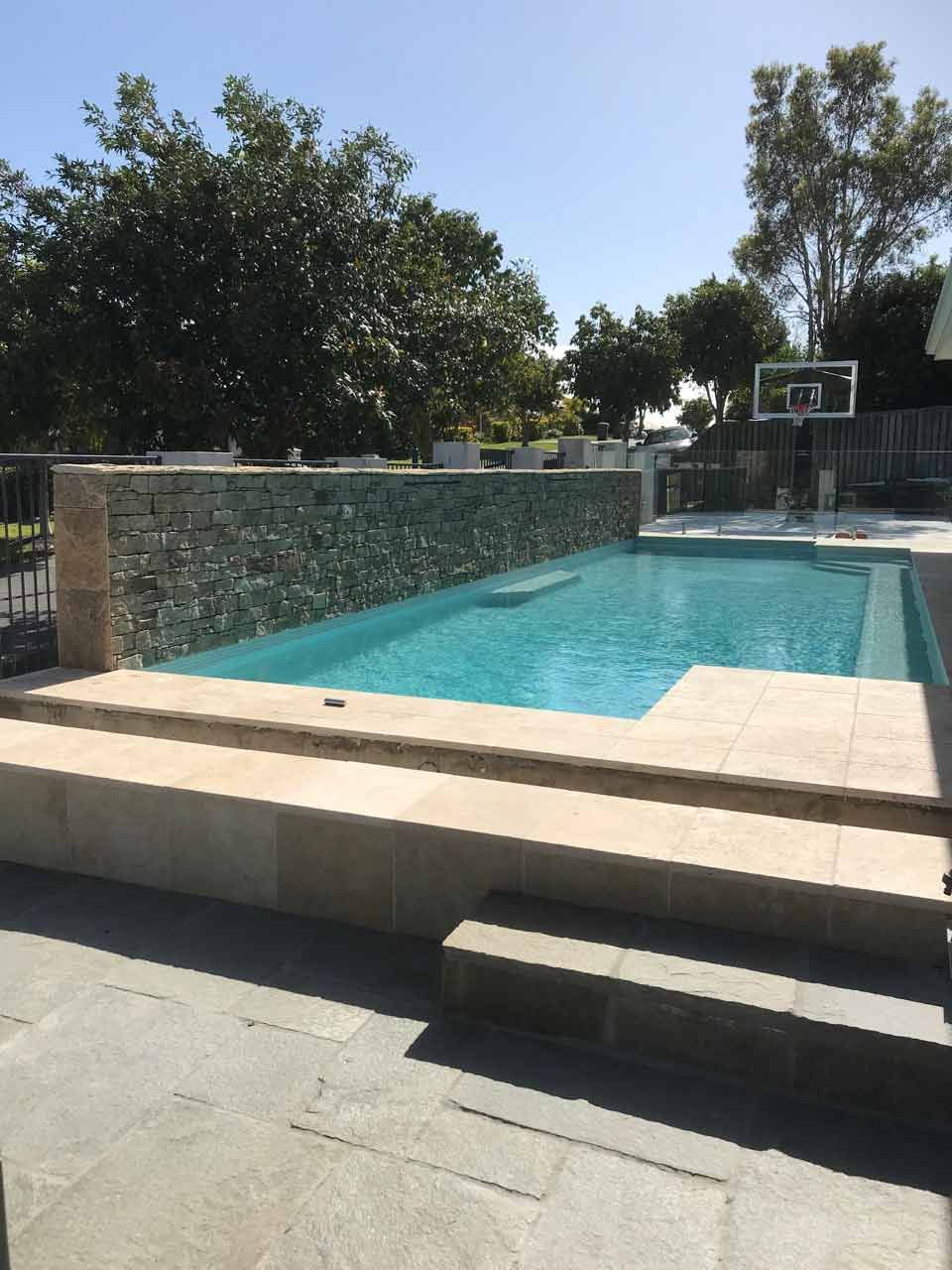 Pool with stone wall and basketball hoop, steps leading into the water, sunny outdoor setting.