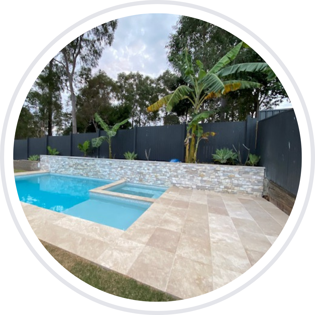Pool area with light-colored stone tiles, blue pool, and a stone wall with greenery against a dark fence.