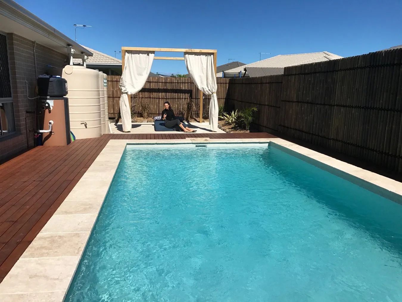 Backyard with pool, wooden deck, gazebo with curtains, bamboo fence, and person seated.