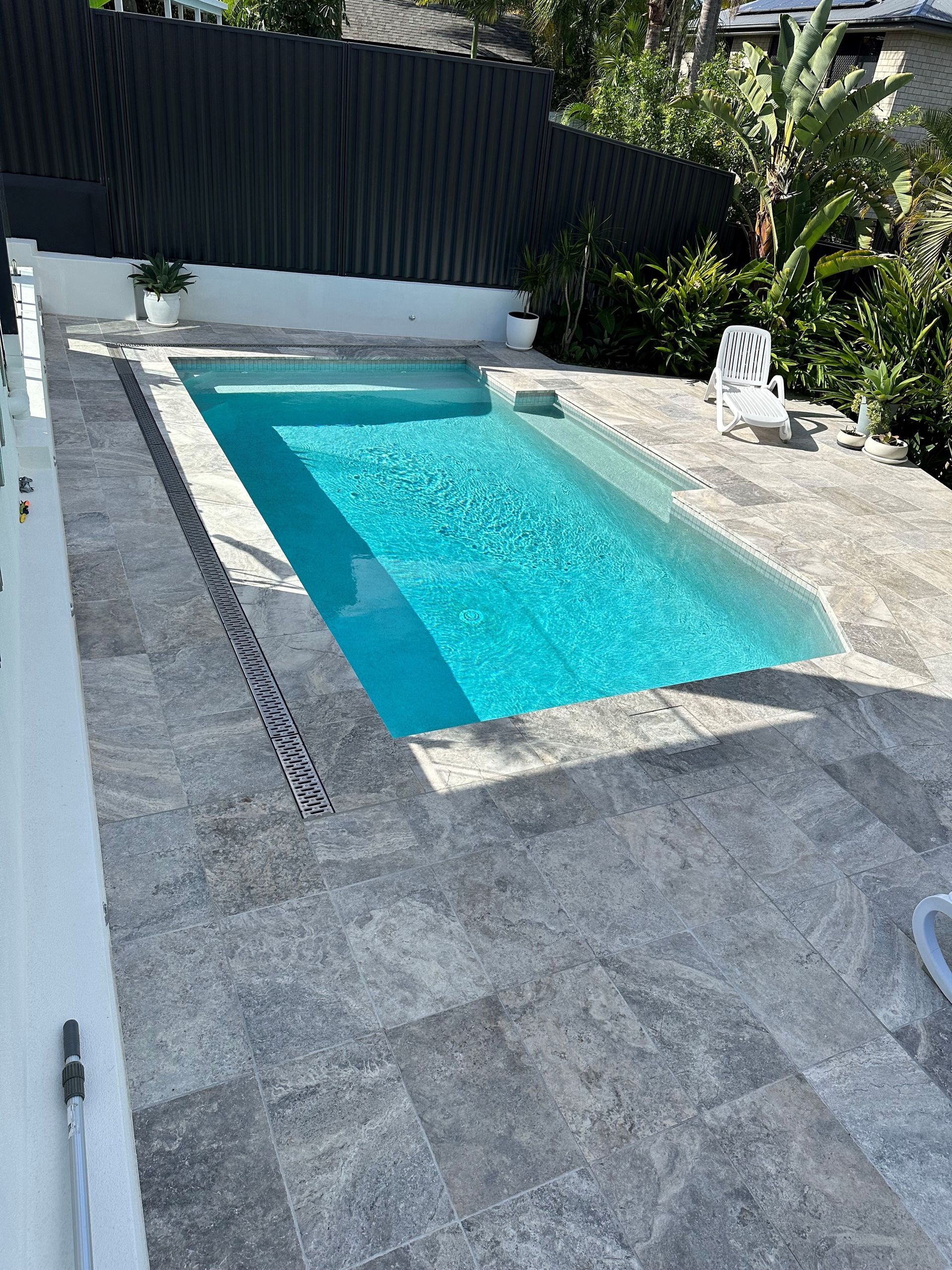 Rectangular turquoise pool surrounded by gray stone patio, white fence, and lush greenery.