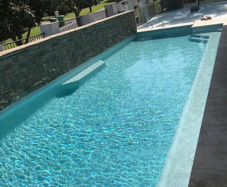 Rectangular swimming pool with clear blue water. A stone wall borders one side; the pool has steps.