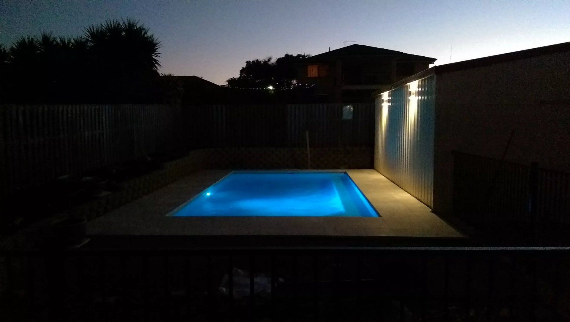 Pool with blue water illuminated at dusk; lit up by a wall with lights; backyard setting.