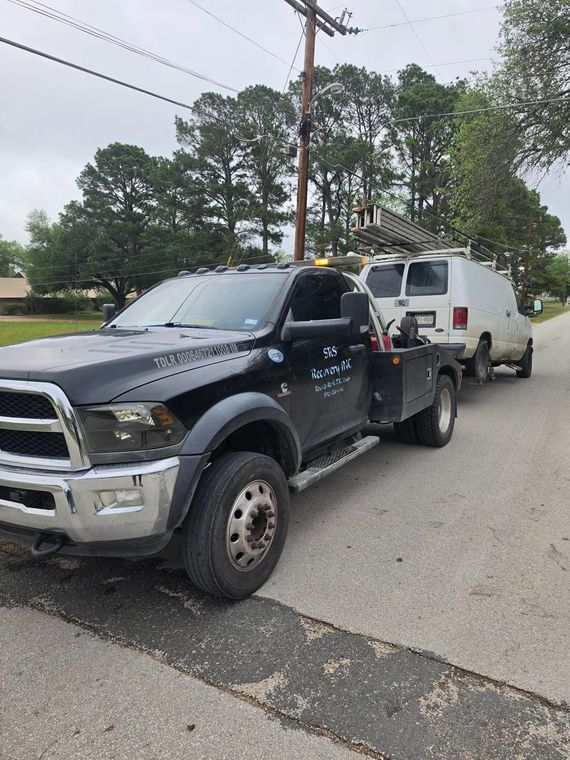 Black tow truck hauling a white van on a residential street under overcast skies