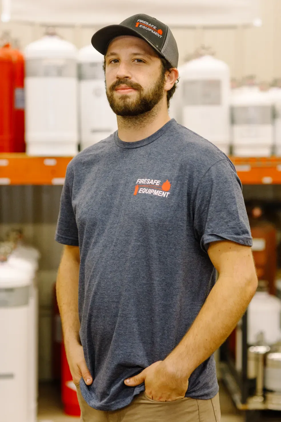 A man wearing a hat and a t-shirt is standing in front of a shelf with fire extinguishers.