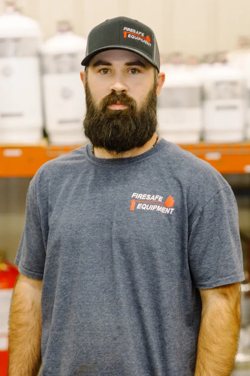 A man with a beard wearing a hat and a t-shirt is standing in front of a shelf.