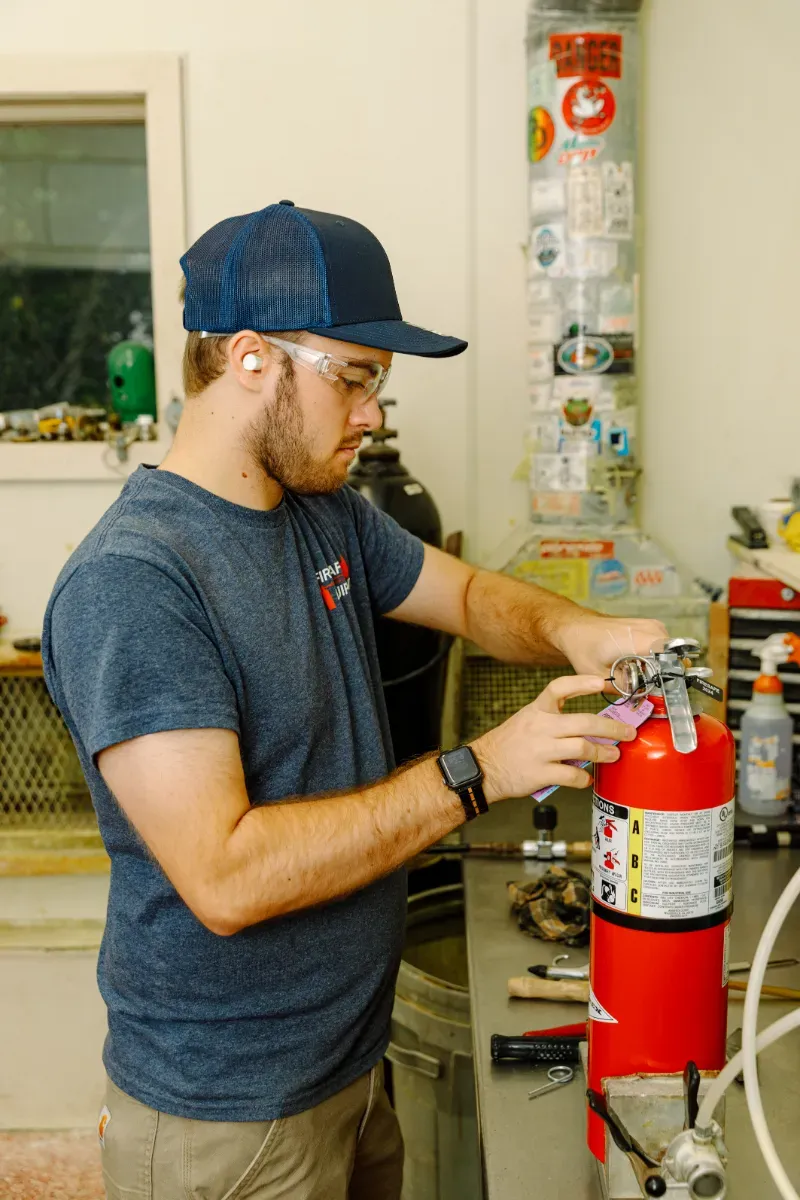 Person in a blue cap adjusts a red fire extinguisher in a workshop.