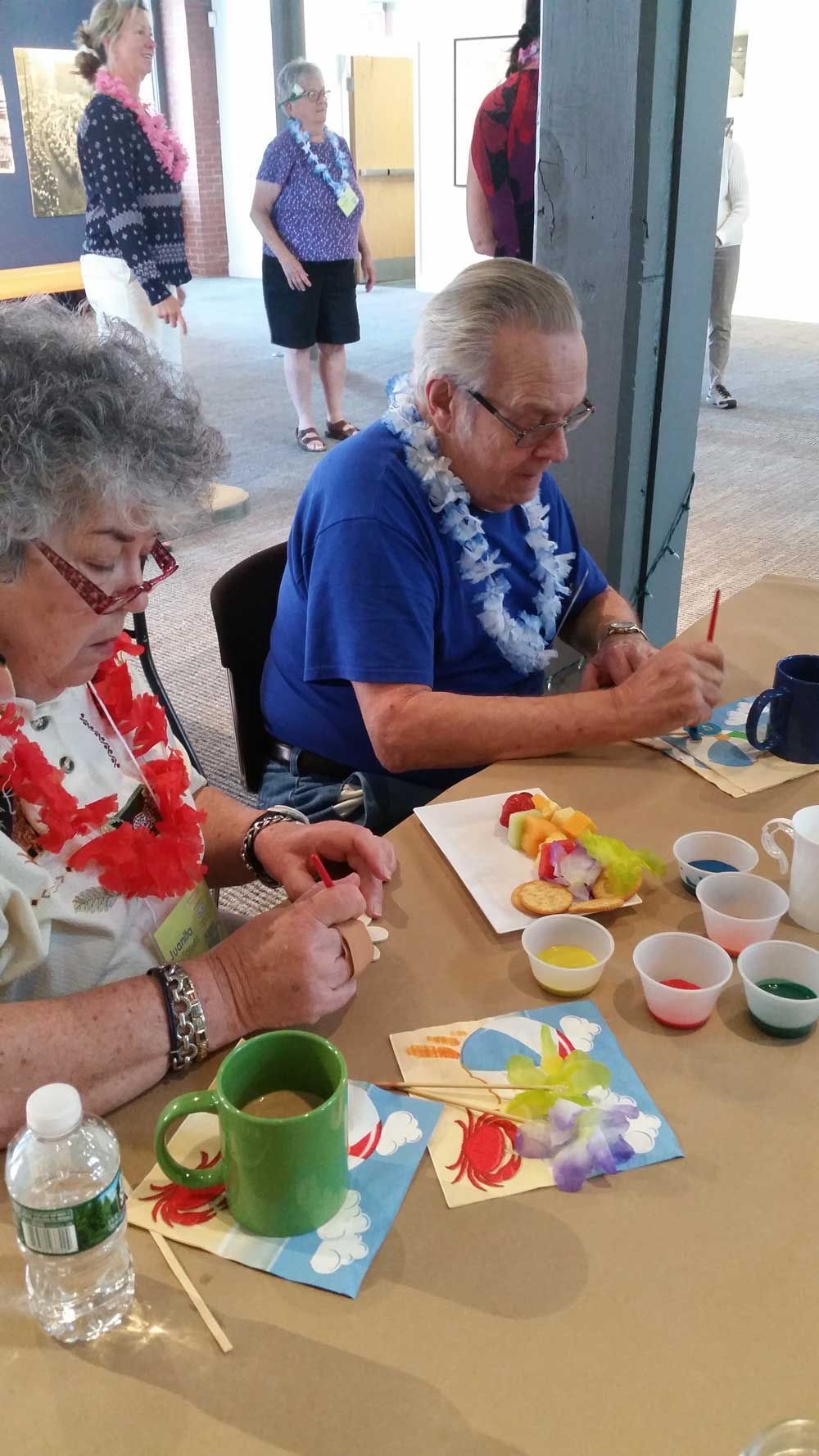 Two people painting at a table with small paint cups, brushes, and colorful paper in a bright indoor space