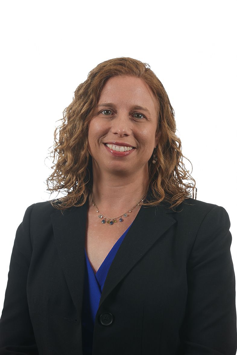 Woman with curly hair in a black blazer over a blue top, smiling against a white background