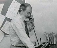 Man with beard on the phone in an office, papers on the wall and desk.