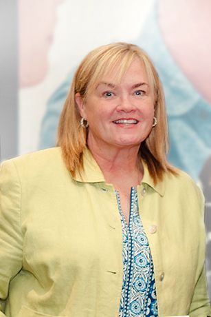 Smiling woman in a light green jacket over a patterned blouse, standing against a soft pastel background
