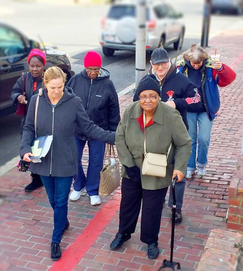 Group of people walking on a brick sidewalk. One uses a cane. Cars are in the background.
