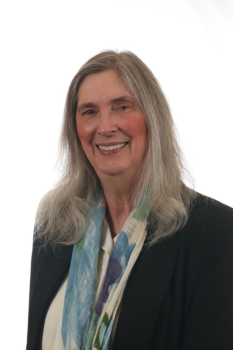Smiling woman with long gray hair in a black blazer and blue patterned scarf against a white background