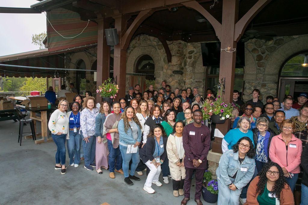 Large group posing outside a stone building with a covered porch, many smiling at the camera.
