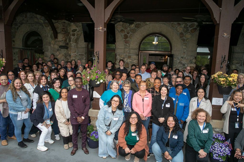 Large group posing indoors under wooden arches, smiling in a stone courtyard with flowers and arched doorways.