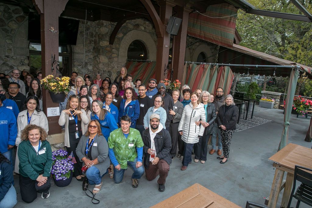 Large group posing outdoors on a covered patio beside a stone building, with benches and greenery nearby
