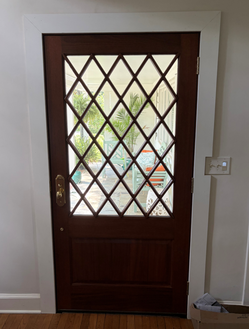 Wooden front door with diamond lattice glass panel and brass handle, opening to an outdoor view.