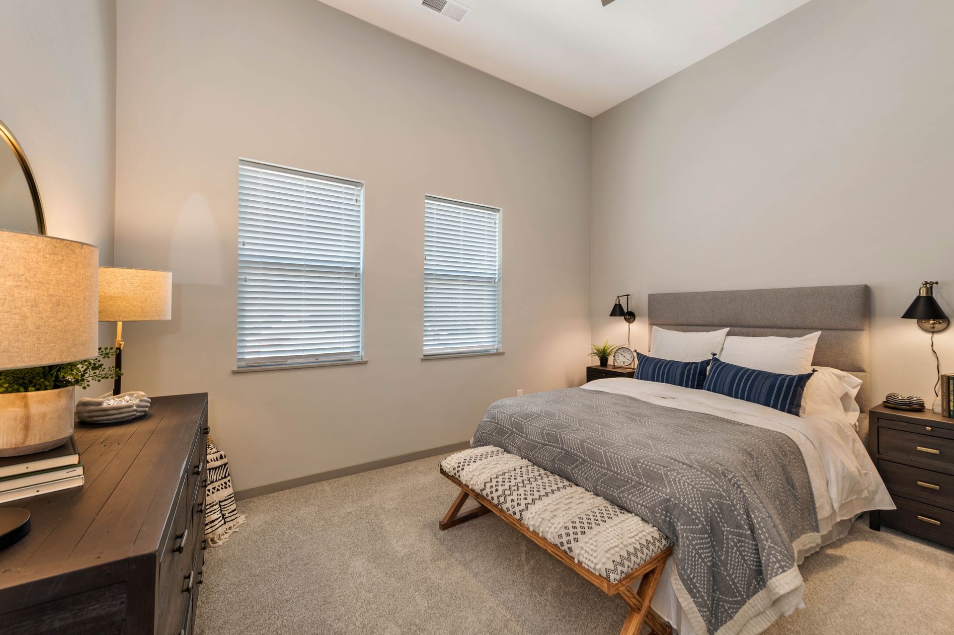 A bedroom with a bed , dresser , lamps and two windows.