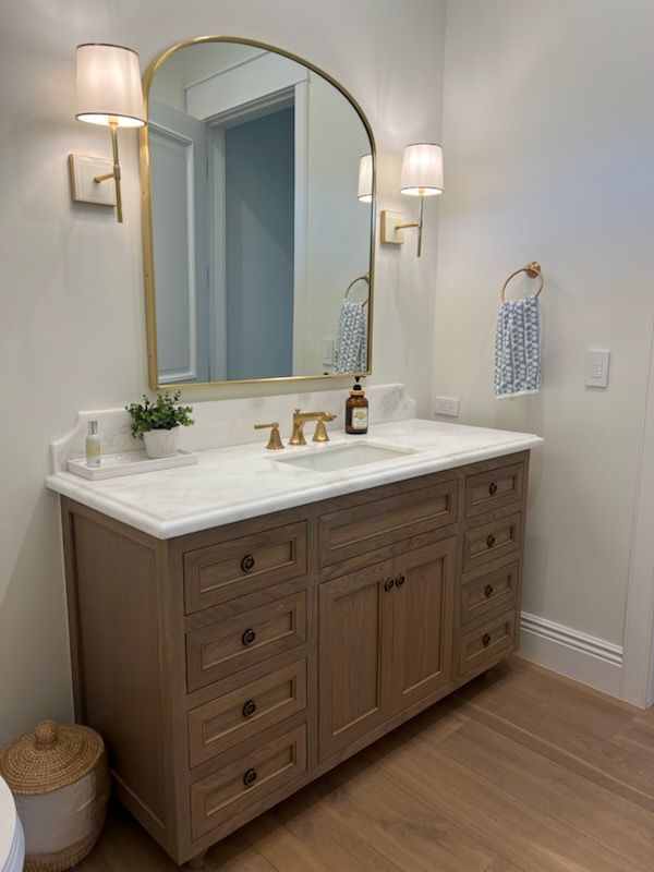 Bright bathroom vanity with wood cabinets, white sink, gold mirror, and wall sconces above hardwood floor.