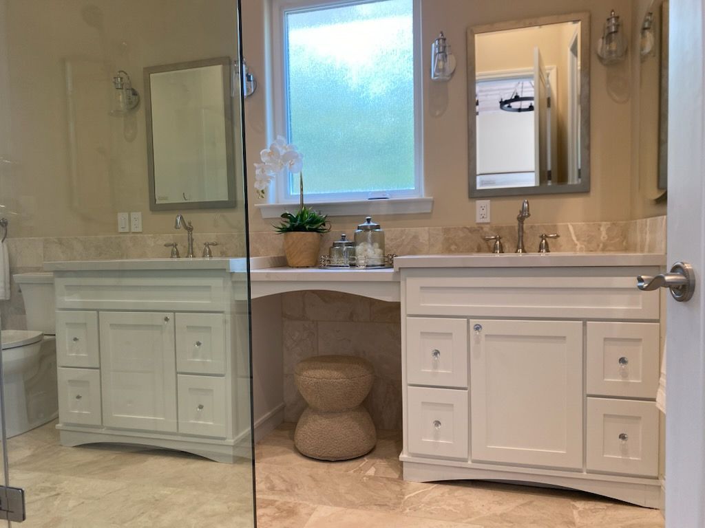Bright beige bathroom with double vanity, mirrors, window, and white cabinets.