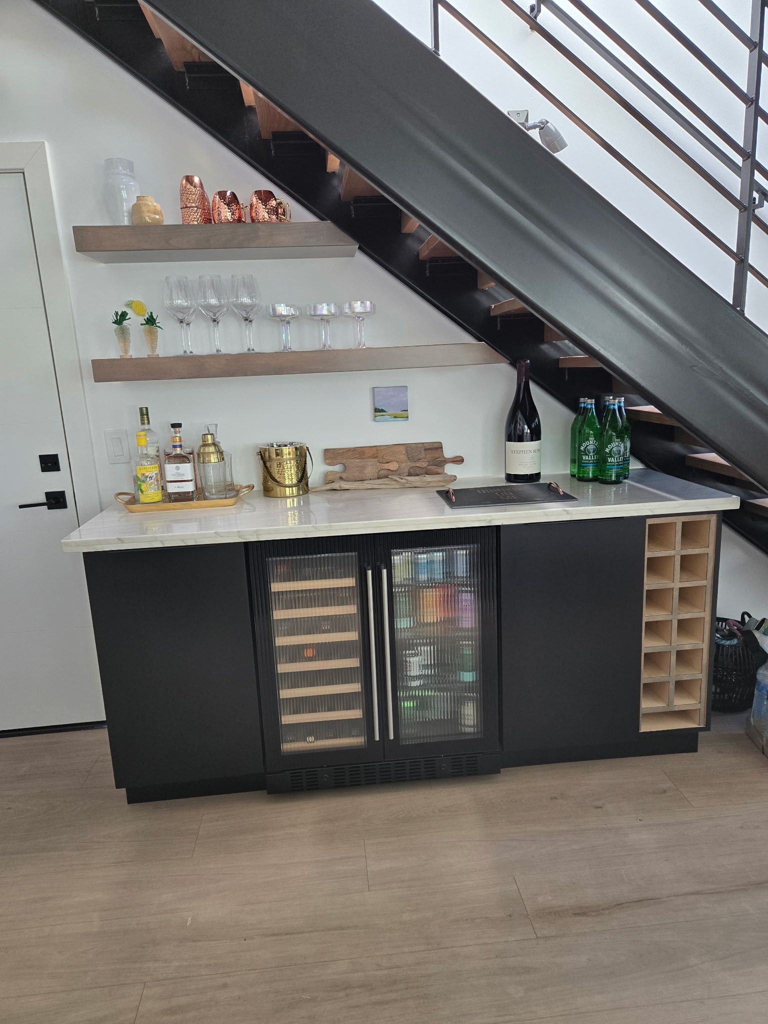 Under-stair home bar with shelves, glass-front fridge, bottles, and wine racks on a wood floor