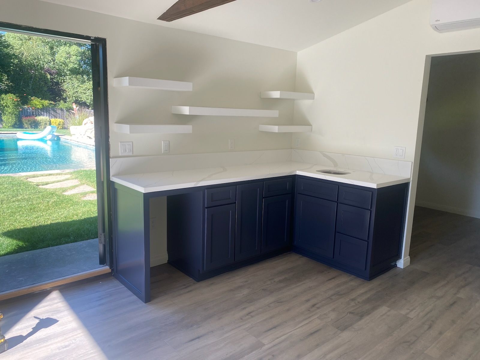 Empty corner room with dark cabinets, white countertops, shelves, and a sliding glass door to a poolside patio