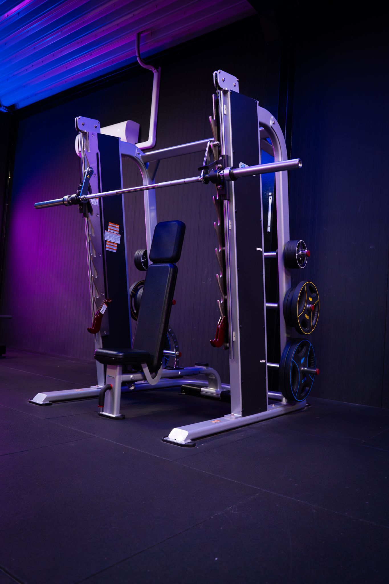 Paars verlichte sportschool met een Smith-machine, een bankje en gewichtsschijven op een donkere vloer.