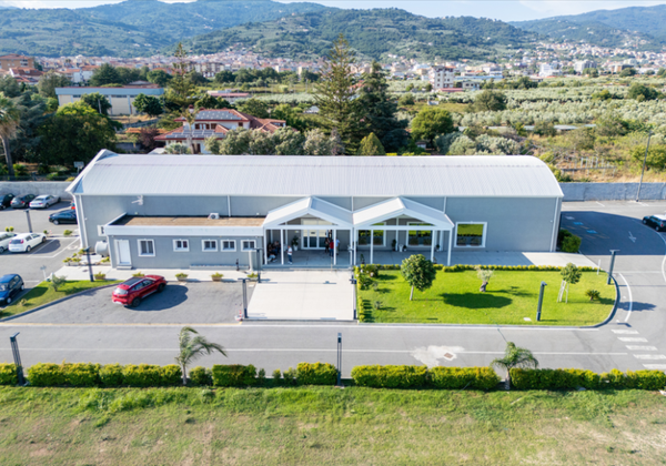 Veduta aerea di un moderno edificio bianco con un parcheggio, una strada e delle colline sullo sfondo.