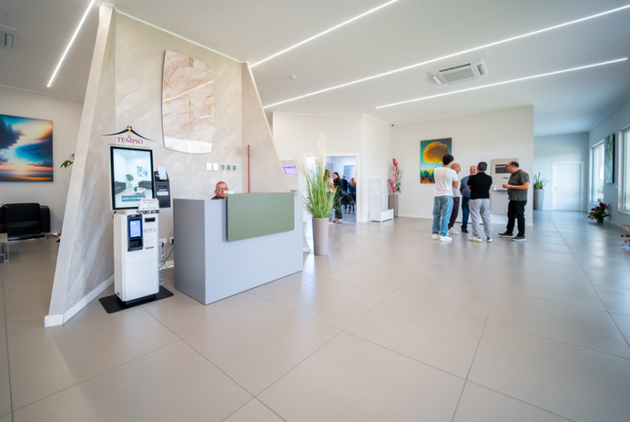 Hall moderna con chiosco self-service, banco reception e un piccolo gruppo di persone che chiacchierano sullo sfondo.