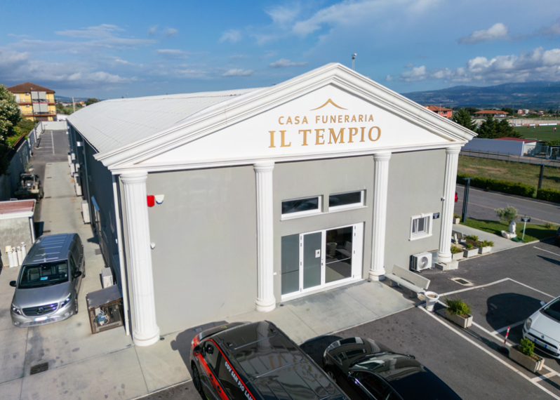 Veduta aerea della Casa Funeraria Il Tempio, un edificio bianco con auto parcheggiate e montagne sullo sfondo.