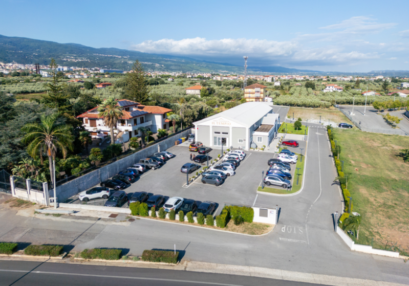 Veduta aerea di un parcheggio adiacente a un edificio commerciale bianco e alle case circostanti, con le montagne sullo sfondo.