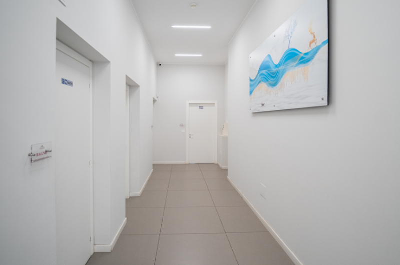 Luminoso corridoio bianco con pavimento in piastrelle beige, porte chiuse e quadro astratto blu alle pareti.
