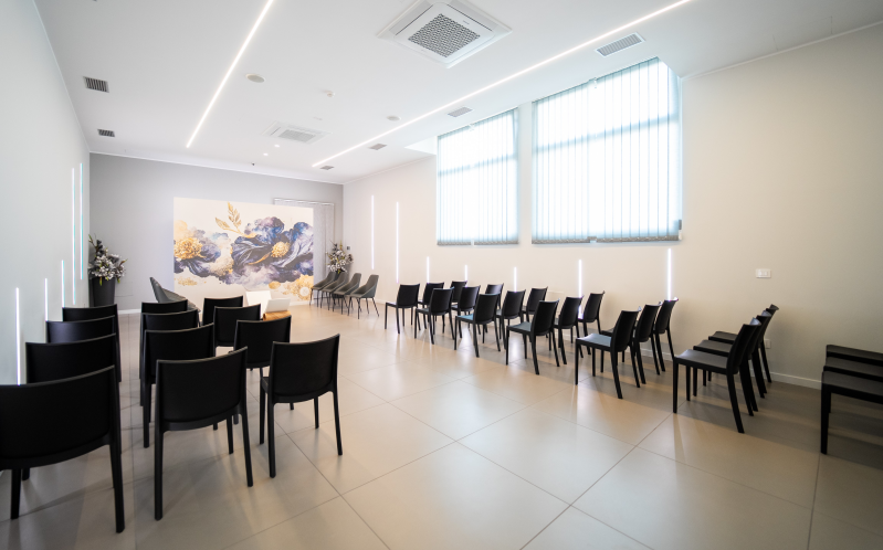 Luminosa sala conferenze con sedie nere, pareti bianche, pavimento piastrellato e grandi finestre, allestita per un evento.