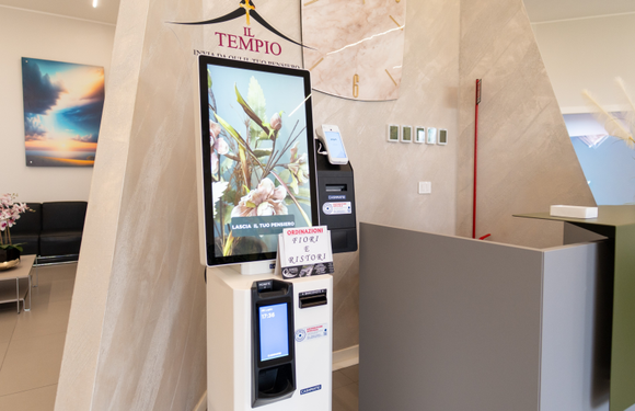 Chiosco per i pagamenti nella hall dell'hotel, con schermo digitale e piccolo cartello, accanto a un bancone della reception beige.