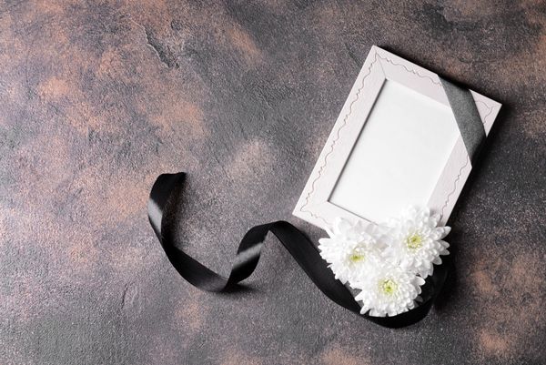 Cornice portafoto bianca con nastro nero e fiori bianchi su sfondo grigio testurizzato.