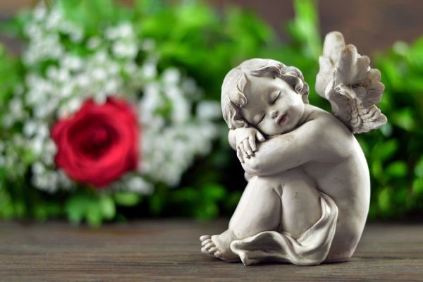 Statuetta di un angelo bianco appoggiata su una superficie di legno, con una rosa rossa e fiori sfocati sullo sfondo.