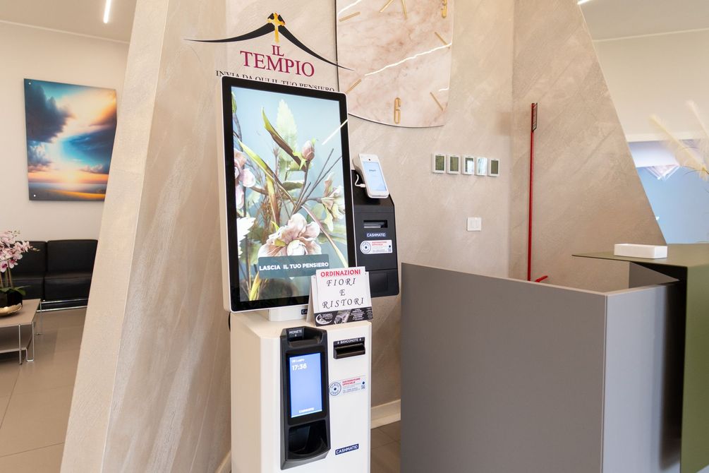 Chiosco per i pagamenti nella hall dell'hotel, con schermo digitale e piccolo cartello, accanto a un bancone della reception beige.
