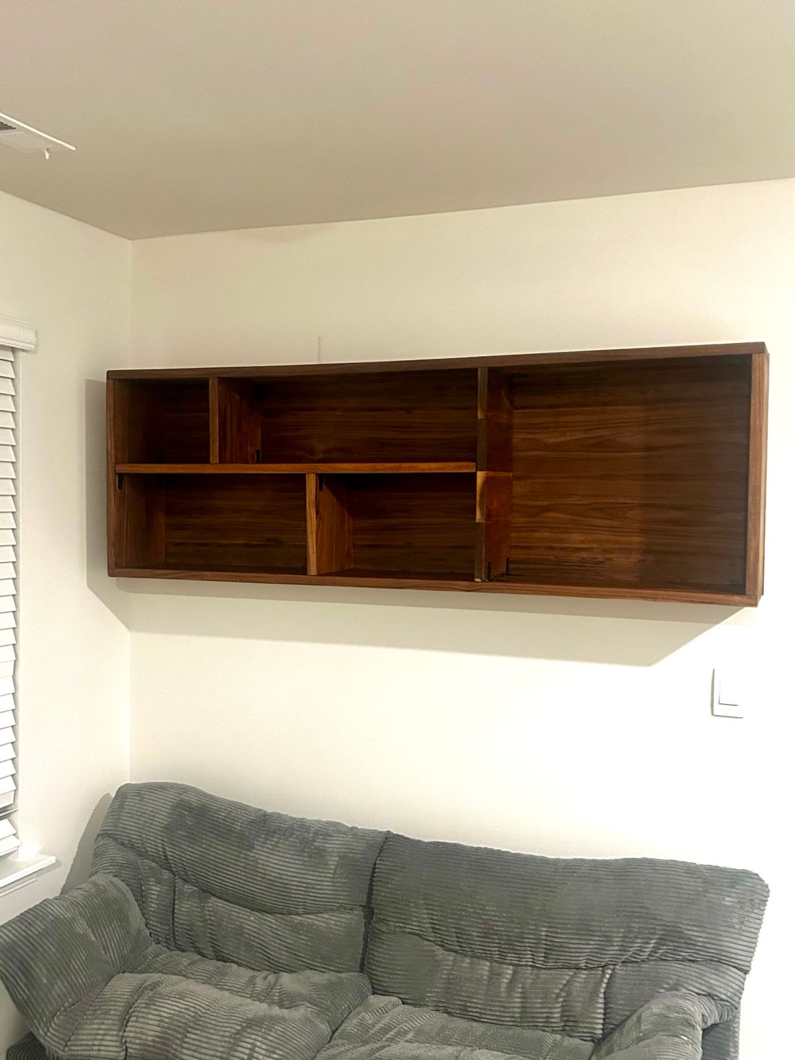 Living room corner with a wall-mounted wooden shelf above a gray couch and window blinds on the left.