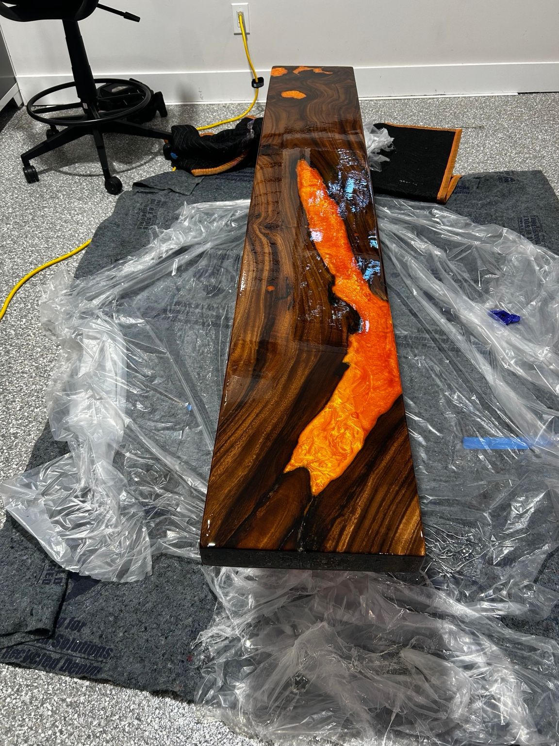 Wood slab with glowing orange resin inlay, drying on a plastic-covered workshop floor.