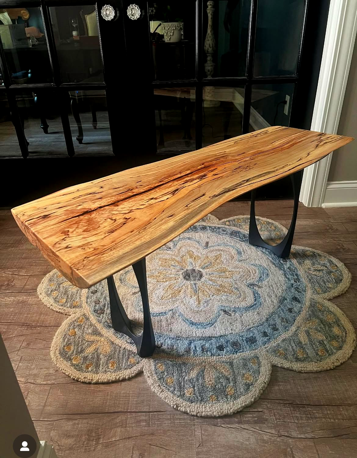 Live edge wood bench on black metal legs, set on a floral rug over wood flooring.