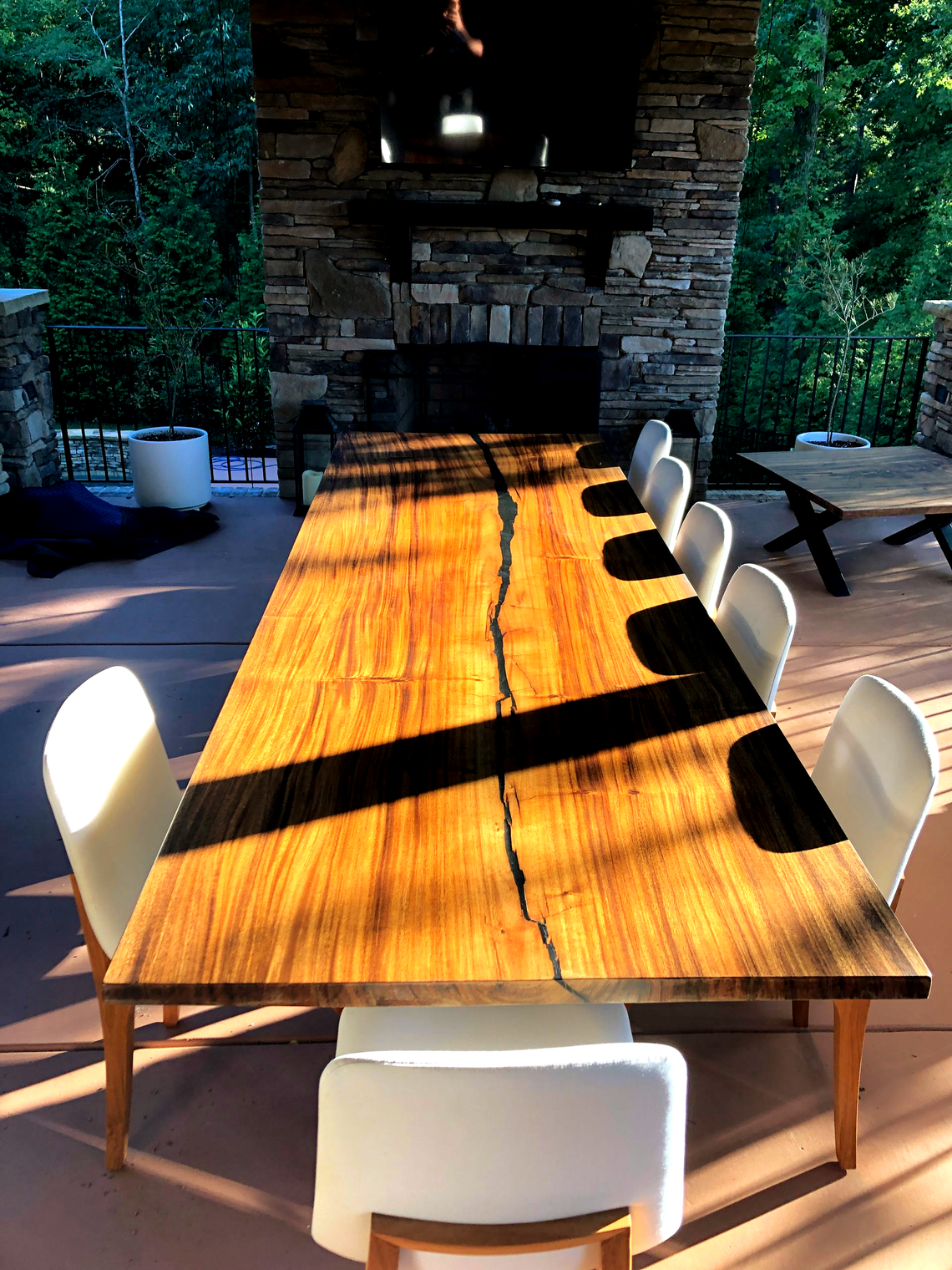 A long, wooden slab table with a dark center seam sits on a patio with white chairs, facing a stone outdoor fireplace.