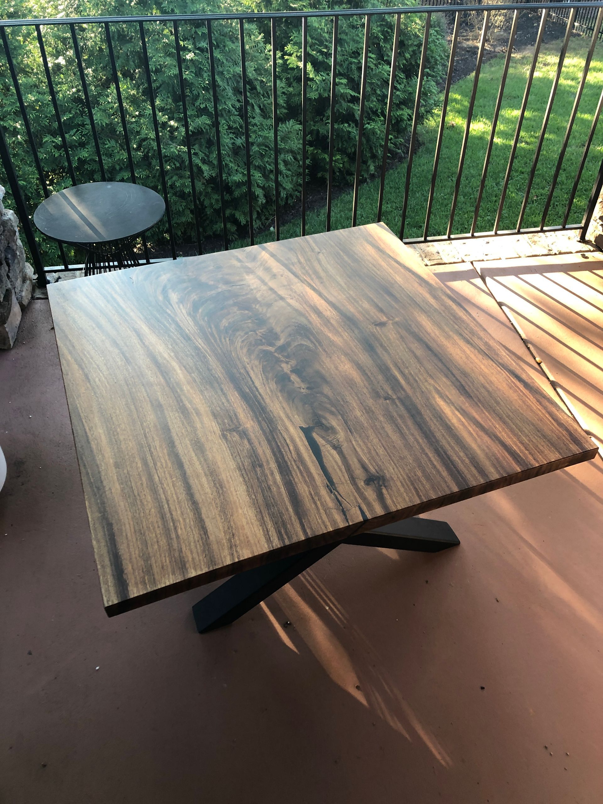 Wooden square table with dark base on a deck, overlooking a green yard.
