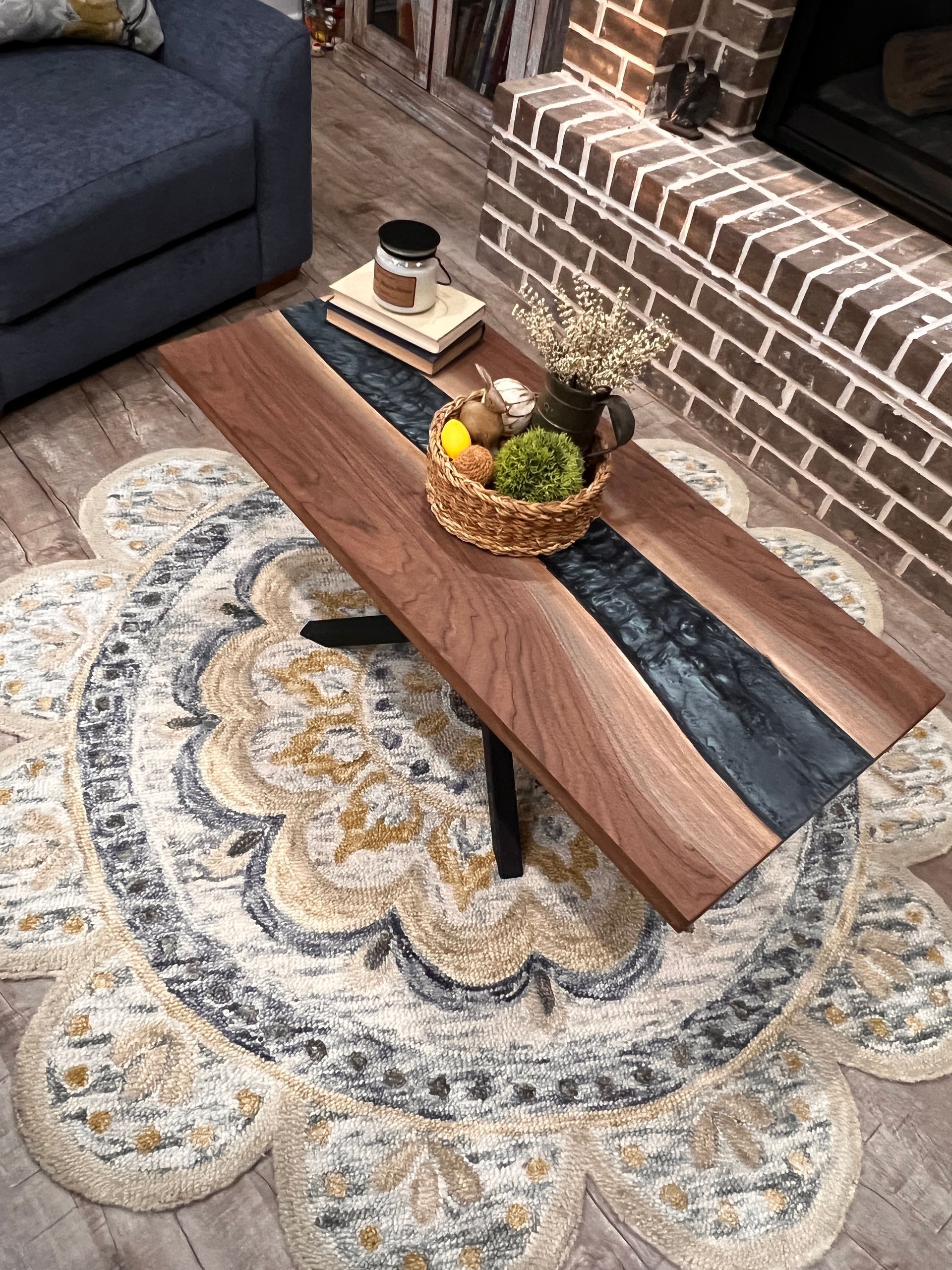 Wooden coffee table with dark epoxy river, basket of fruit, rug, and brick fireplace.